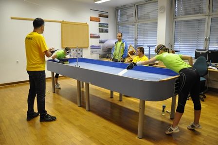 Several people in colorful t-shirts stand around a long table playing a game called Showdown