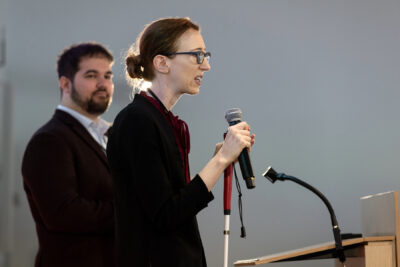 Shiri Azenkot holding white cane and microphone and making address from podium. Dylan Fox stands beside.