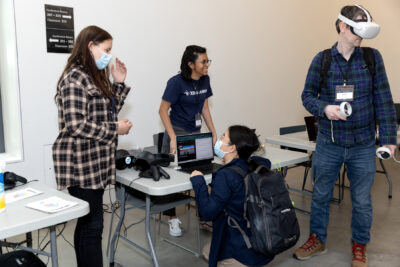 During demos, one person tries a VR headset out while another crouches in front of a laptop. Smiling volunteers assist them.