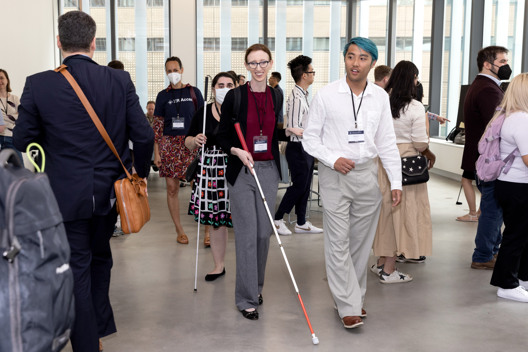 Shiri Azenkot, a thin red-headed woman with a white cane, walks through a crowded demo hall at the 2023 XR Access Symposium