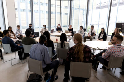 Liv Erickson, a woman in a black dress, leads a large breakout group, multiple tables surrounded by seated attendees.