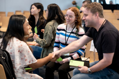 Two people shake hands while a third smiles and looks on.