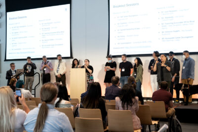 Danielle Montour stands behind the podium holding her white cane, with the breakout session leaders arrayed behind her. Viewed from the front, with the breakout schedule displayed onscreen. There are 12 breakout leaders, one of whom holds the lead of a guide dog.