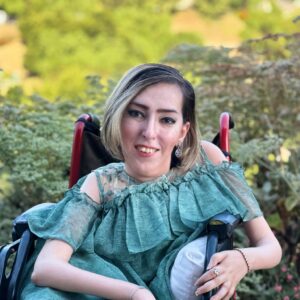 Headshot of Atieh Taheri, a woman with streaked blonde hair and wearing a teal dress seated in a wheelchair.