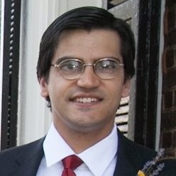 Headshot of Agustya Mehta, a dark-skinned man with neatly combed hair and thin wire glasses wearing a suit and tie.