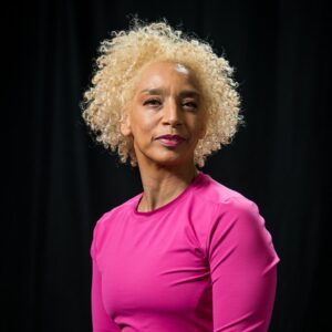  A headshot of Alice Sheppard; she is a multiracial Black woman with coffee-colored skin and platinum blond short curly hair, she wears a hot pink top. Alice regards the camera with a slight smile. Photo by Cherylynn Tsushima.
