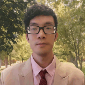 Headshot of Khang Dang, a Vietnamese man with short, wavy black hair and glasses. He is wearing a light beige suit jacket, a pale pink dress shirt, and a textured red tie. He has a calm, neutral expression. The background shows a sunny, tree-filled park with green and golden foliage.