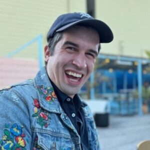 Headshot of Matthew Bambach, a white man with a big smile wearing a baseball cap and jean jacket embroidered with flowers.
