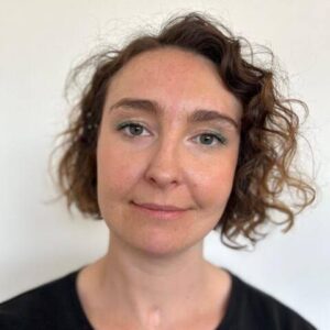 A headshot of Meg Fozzard, a white woman with short curly hair in a side parting. The left side of her face is bathed in pink light and the right hand side in a bluey/purple. She has a neutral expression. 