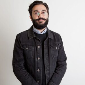 Headshot of Milad Mozari, a brown-skinned man with a neat beard and circular gold-rimmed glasses wearing a black jacket over a collared shirt and sweater.