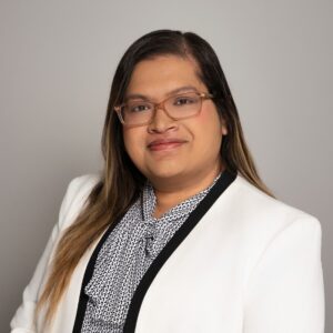 Headshot of Rupsha Mutsuddi, a brown woman with long dark hair wearing a white suit and a soft smile against a grey backdrop