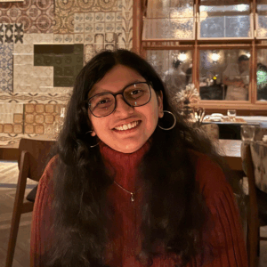 Headshot of Mrunmai Abhyankar, a light skinned woman with long curly hair and glasses, wearing a red sweater, smiling in an indoor setting with patterned walls in the background. 
