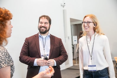 XR Access leaders Dylan Fox, a white man with short brown hair and beard wearing a purple suit jacket; and Shiri Azenkot, a white woman with light brown hair wearing a white blouse; smile as they chat with an attendee.