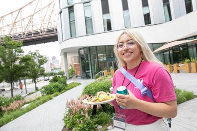 Jazmin Cano of Owlchemy Labs, a light-skinned woman with long platinum blonde hair wearing a pink shirt, smiles as she holds up a plate of tacos outside the venue. The Queensboro Bridge extends overhead in the background.