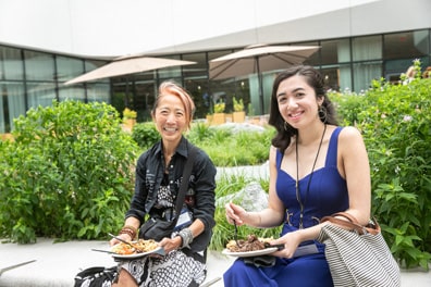 Two women smile at the camera as they enjoy their lunch, surrounded by bright green bushes in the space outside the venue.