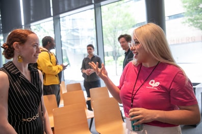 Jazmin Cano and Jazmin Collins chat during a break; the latter is wearing earrings resembling a yellow Nintendo Game Boy. Three more attendees form another small group in the background.
