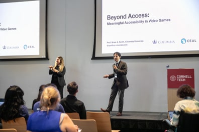 Brian Smith, a light-skinned man with curly brown hair wearing a black jacket, strikes a jaunty pose as he presents Beyond Access: Meaningful Accessibility in Video Games.