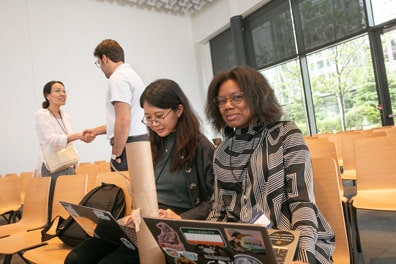 Judite Blanc sits next to another attendee as they both work on laptops, Judite peeking up to smile at the camera. Behind them, the two other attendees shake hands.