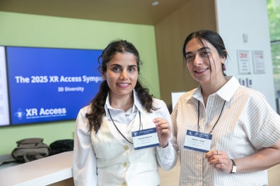 Attendees Maryam Bigonah and Niloofar Sayadi, both olive-skinned women with long black hair, smile and hold up their nametags in front of a monitor proudly displaying the Symposium title slide.