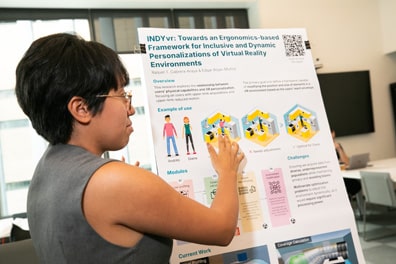 Raquel T. Cabrera-Araya, a brown-skinned woman with short black hair and glasses, points out a feature on her poster: Indy VR, Towards an Ergonomics-Based Framework for Inclusive and Dynamic Personalizations of Virtual Reality Environments.