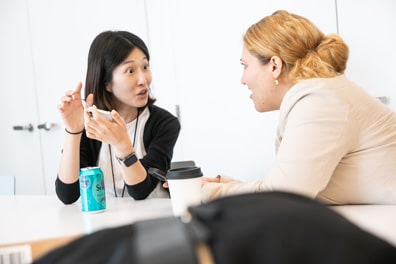 Two women engage in a discussion, one with eyes wide as she excitedly referencing something on her smartphone.