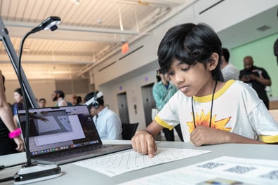 A young dark-skinned boy wearing a yellow-rimmed T shirt feels a tactile display at one of the demo tables.