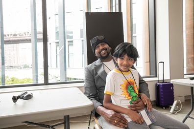 The boy sits in his fathers lap next to one of the demo tables, both smiling at the camera.