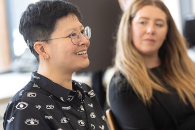 Closeup of a smiling attendee, a short-haired Asian woman with circular glasses, several ear rings, and a black collared shirt featuring open and closed white eyes.