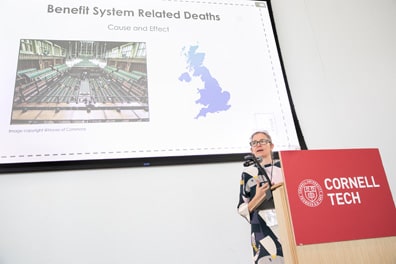 Sacha Wares, a white woman with sideswept salt-and-pepper hair wearing a dress with abstract designs, gestures emphatically during her presentation “Accessibility Innovations for Extended Reality Drama and Documentary.” Behind her, a slide titled “Benefit System Related Deaths: Cause and Effect” shows a mostly empty legislative chamber and the outline of Britain.