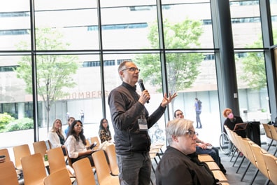 An audience member stands and asks a question into the microphone, his fingers lightly extended in an open-palm gesture. Behind him, the broad glass walls of the Verizon Center show some greenery and the Bloomberg building beyond.
