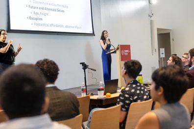 Ria Gualano, a multiracial woman with long, dark brown hair, presents about Designing Disability-Inclusive Avatars to a rapt audience. Her slide mentions Future and Alternative Selves, and Escapism.