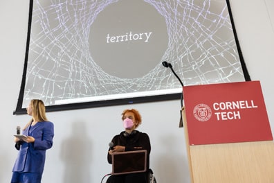Kiira Benz and Alice Sheppard proudly present video from their film Territory. On screen, the title is shown on a black starry sky, surrounded by rings of encircling barbed wire. Kiira is a light-skinned woman with long blonde hair wearing a blue suit; Alice is a multiracial Black woman with coffee-colored skin and platinum blond short curly hair in black clothes seated in a wheelchair.