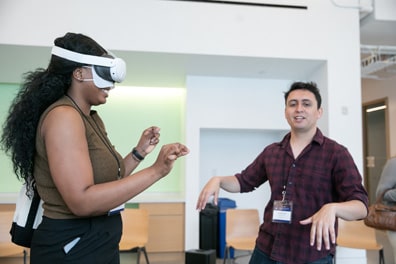 A participant wearing a VR headset smiles widely as she pinches the air. Next to her, an exhibitor stands mid-gesture, with arms spread and wrists pointing down. The participant is a dark-skinned woman with long curly black hair; the exhibitor is a lighter-skinned man with short dark hair wearing a plaid long-sleeved shirt rolled up to his elbows.