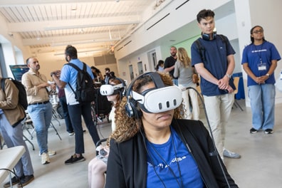 Two attendees experience a virtual reality exhibit in the exhibition hall, which is full of people engaged in conversation.
