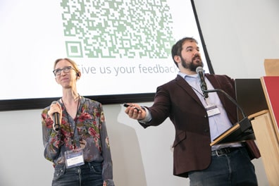 Shiri Azenkot and Dylan Fox, the leaders of XR Access, present from the podium at the closing of the 2025 Symposium, Dylan with his arms wide. Dylan Fox is a white man with short brown hair and beard wearing a purple suit jacket; and Shiri Azenkot is a white woman with light brown hair wearing a grey shirt with flower patterns.