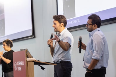 Anis Idrizovic, a white man with wavy brown hair, stands next to copresenter MD Mamunur Rashid, a man with light brown skin, short hair, and a close-cut goatee, wearing black-rimmed glasses. Dressed in matching light blue shirts and navy pants, they share about Augmented Reality Navigation System for Wheelchair Users.
