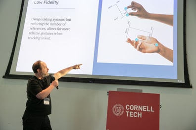 Justin Berry, a balding white man with salt-and-pepper beard, points up at a slide from his presentation Lemmings: Tools For Accessible Gestures. In it, blue dots track distances between various parts of hands, such as the typical pointer finger and thumb pinch, or wrist-to-middle finger.