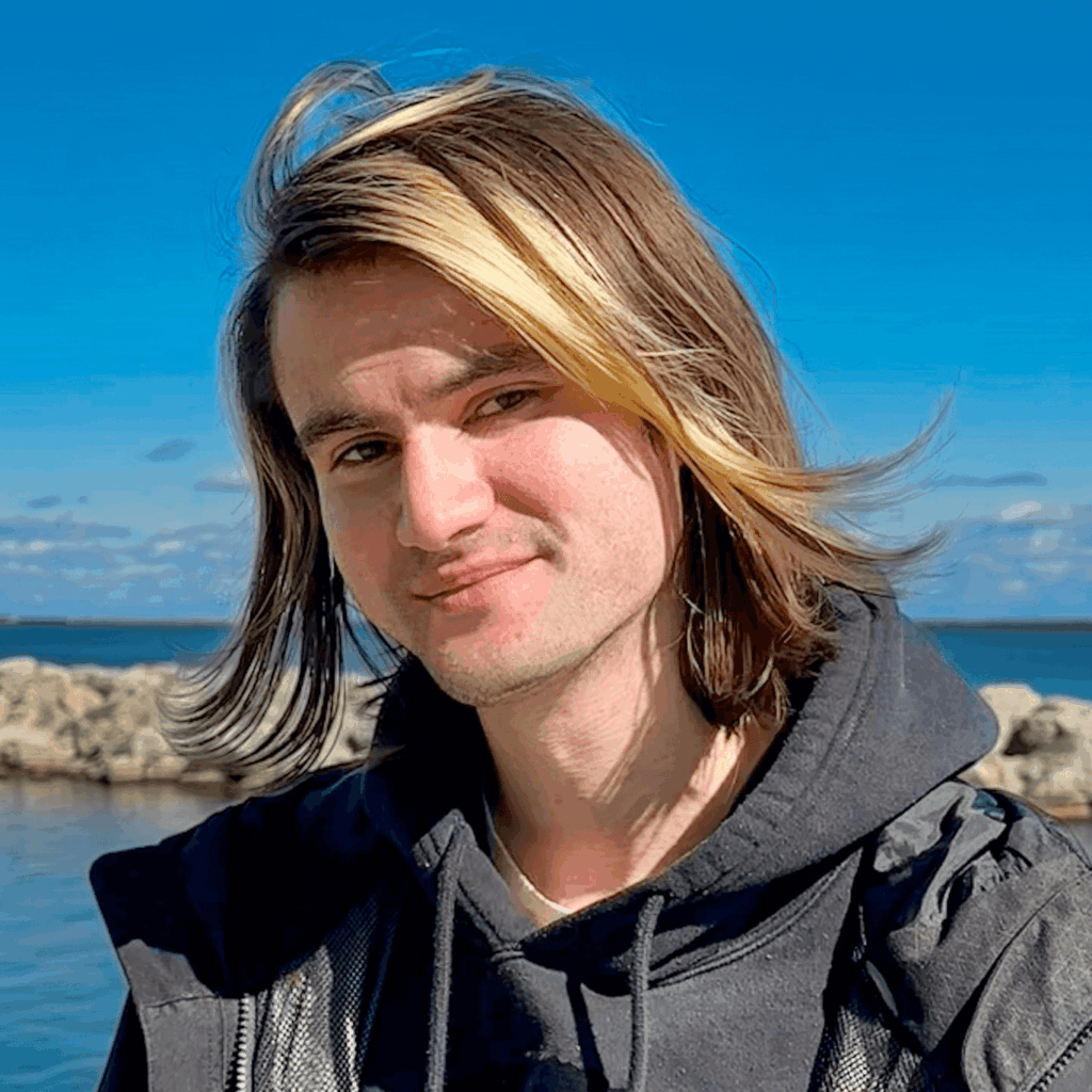 Headshot of Daniel Killough, a light-skinned male-presenting person with long brown hair including gold streaks. He is wearing a black hoodie standing outside. The background contains a blue sky with sparse clouds and a blue lake separated by a line of rocks. 