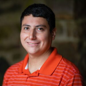Headshot of Brendan David-John, a male in an orange polo and dark hair smiling at the camera while standing against a rock wall.
