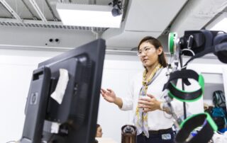Momona Yamagami, an east asian woman, gestures in her lab from behind a monitor and sensor bands in the foreground.
