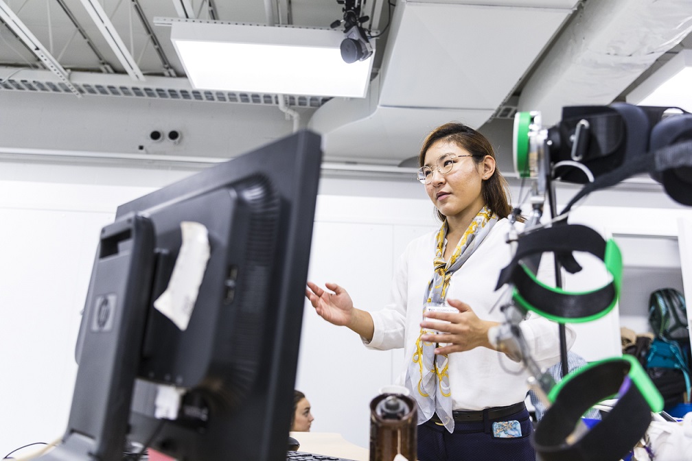 Momona Yamagami, an east asian woman, gestures in her lab from behind a monitor and sensor bands in the foreground.