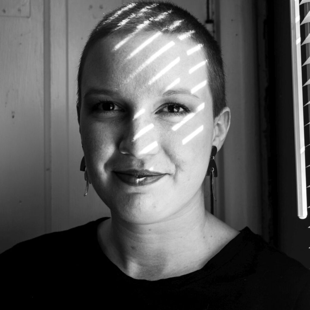 Black and white photo of Laura in a black dress with her head shaved and wearing dangly clay and metal earrings. She is smiling softly and there are stripes of sun falling across her face from the window next to her.